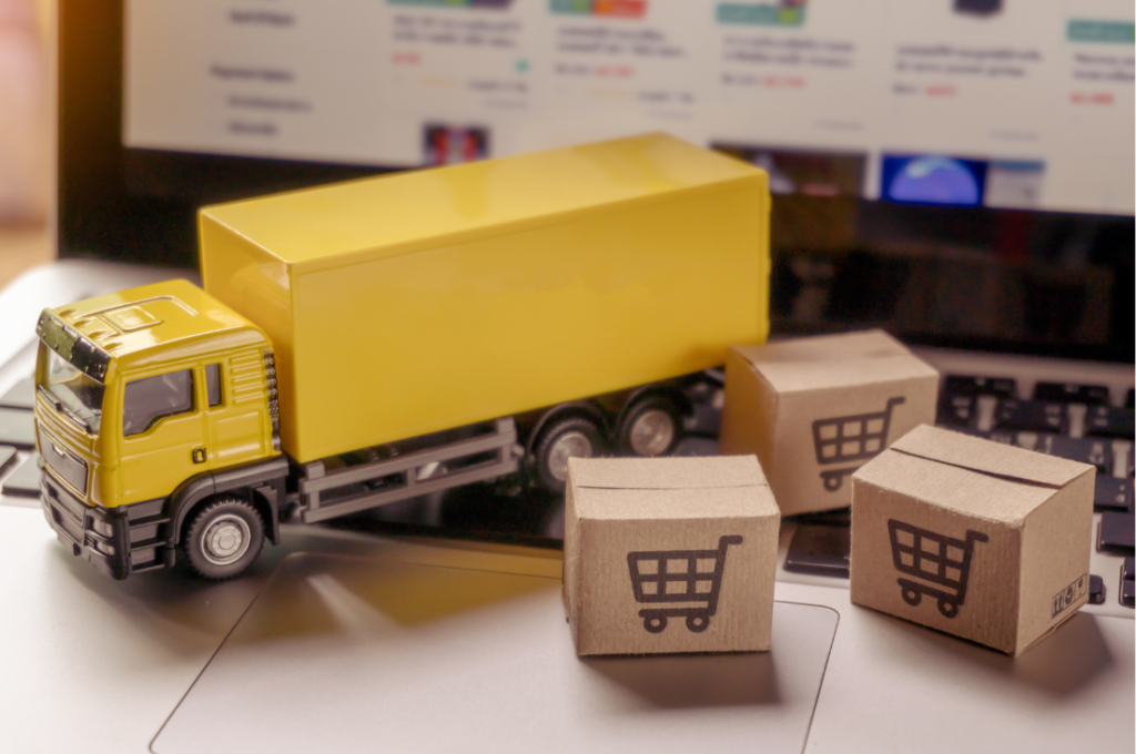 Imagem de um caminhão amarelo em miniatura ao lado de pequenas caixas de papelão com ícones de carrinho de compras, posicionados sobre um teclado de notebook, representando o impacto da logística no e-commerce.