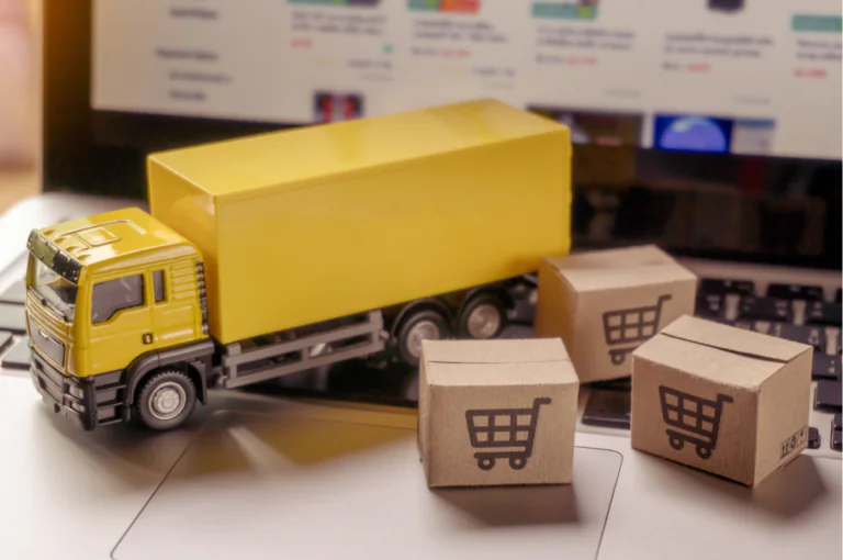 Imagem de um caminhão amarelo em miniatura ao lado de pequenas caixas de papelão com ícones de carrinho de compras, posicionados sobre um teclado de notebook, representando o impacto da logística no e-commerce.